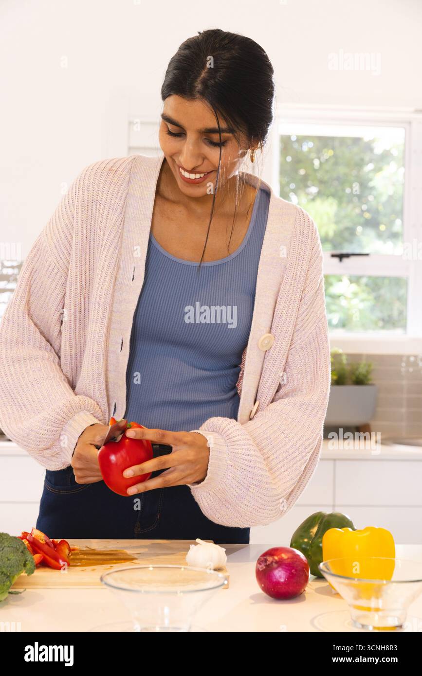 Donna asiatica che affetta peperone rosso sul tagliere con broccoli e cipolla nella cucina moderna Foto Stock