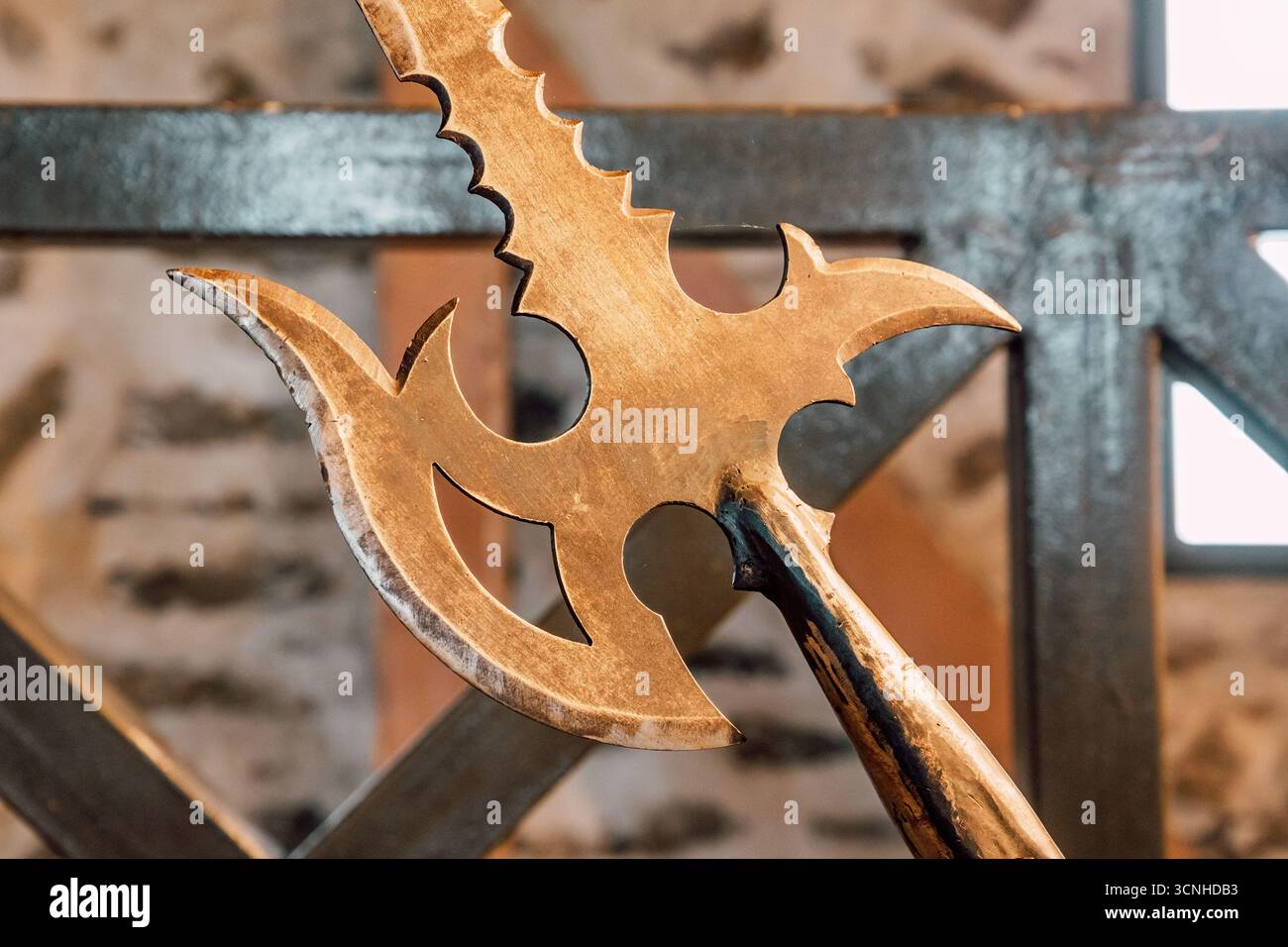 Primo piano di un'alabarda dorata con lame affilate, che evocano fantasia e battaglie antiche, collocata vicino a una recinzione metallica su uno sfondo sfocato Foto Stock