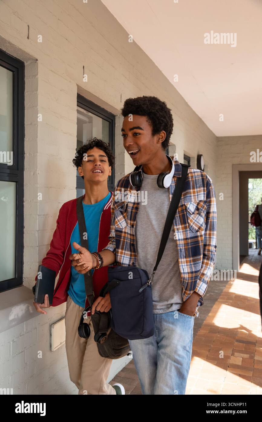 Amici studenti di sesso maschile che camminano e chiacchierano sul passaggio pedonale del campus tenendo in mano tablet e borsa Foto Stock