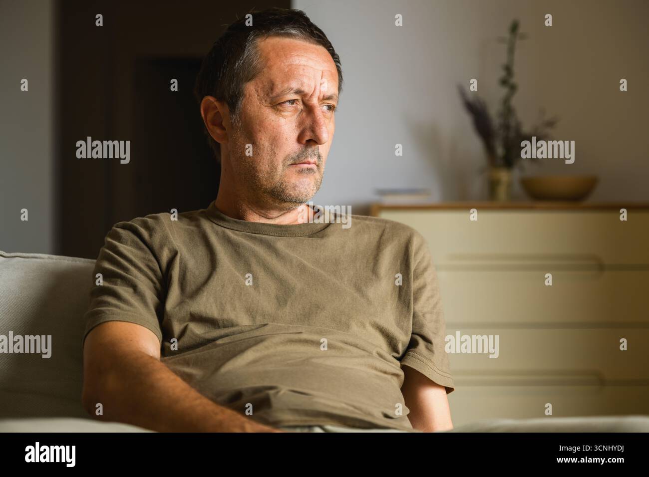 Uomo adulto maturo seduto sul divano di casa, guardando fuori dalla finestra con un'espressione premurosa, simboleggiando la riflessione, il processo decisionale e la contemplazione Foto Stock