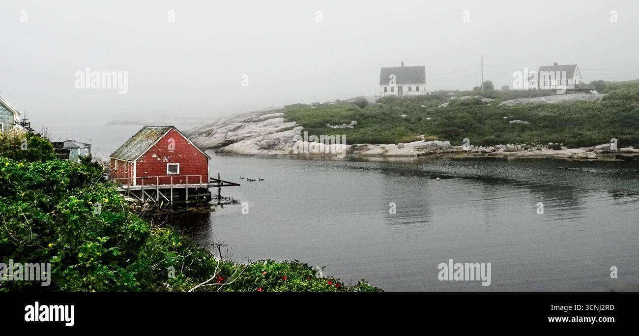 Una tranquilla scena costiera a Peggy's Cove, la nuova Scozia cattura una casetta rossa annidata dall'acqua, circondata dalla nebbia. Le vecchie case punteggiano il paesaggio Foto Stock