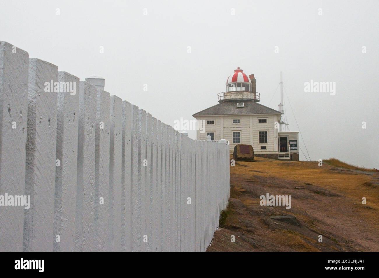 Lo storico faro di Cape Spear è avvolto dalla nebbia, con una suggestiva cupola rossa e pareti imbiancate su una costa rocciosa. Una recinzione bianca conduce verso Foto Stock