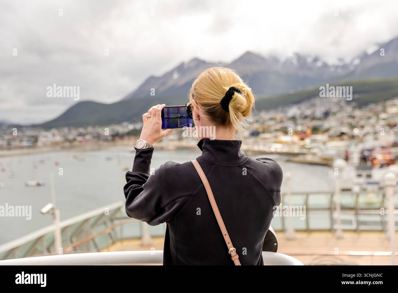 Una donna bionda cattura il suggestivo porto e lo skyline montuoso di Ushuaia con il suo smartphone dal ponte di una nave da crociera. Foto Stock
