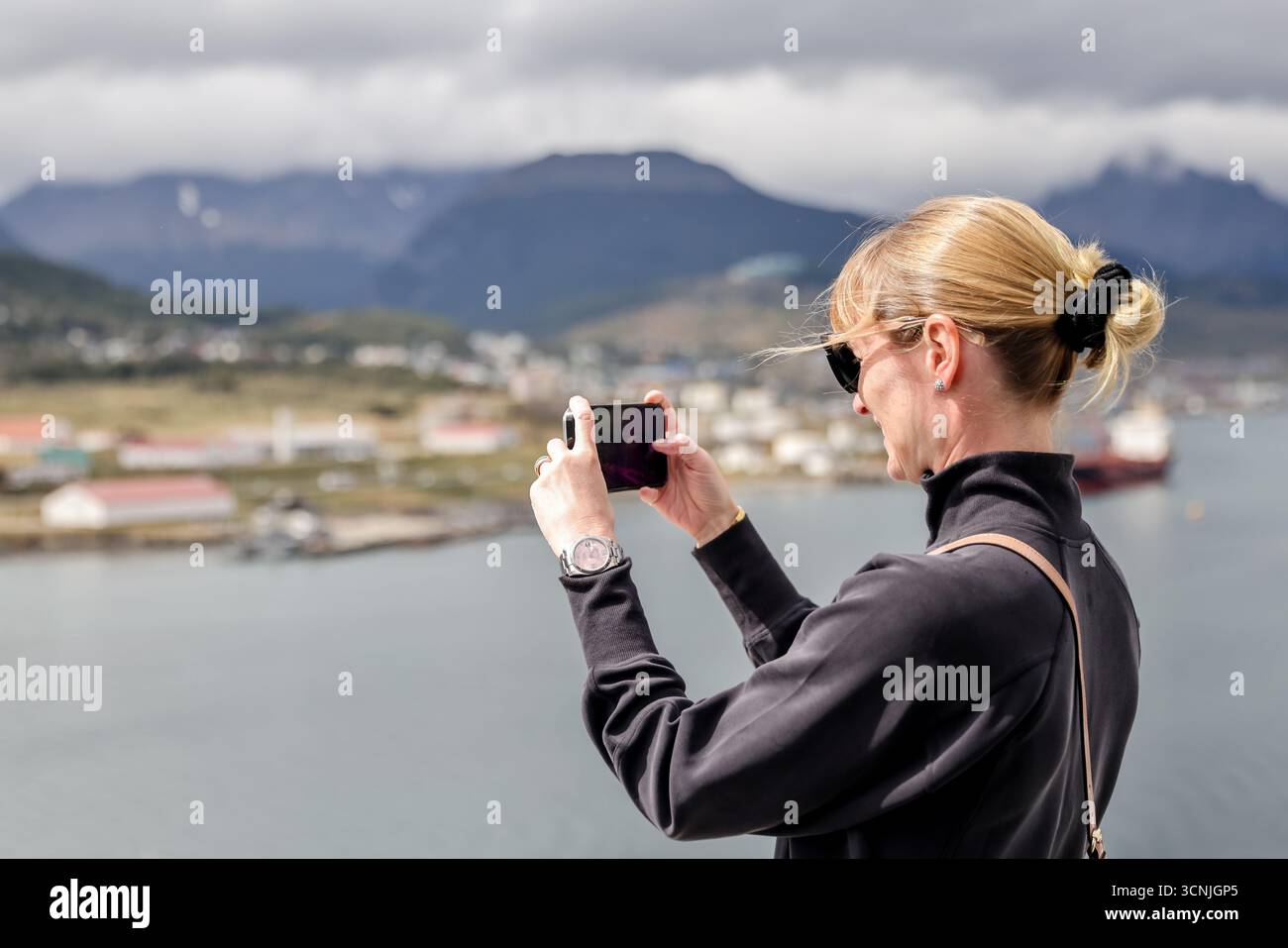 Una donna bionda cattura il suggestivo porto e lo skyline montuoso di Ushuaia con il suo smartphone dal ponte di una nave da crociera. Foto Stock