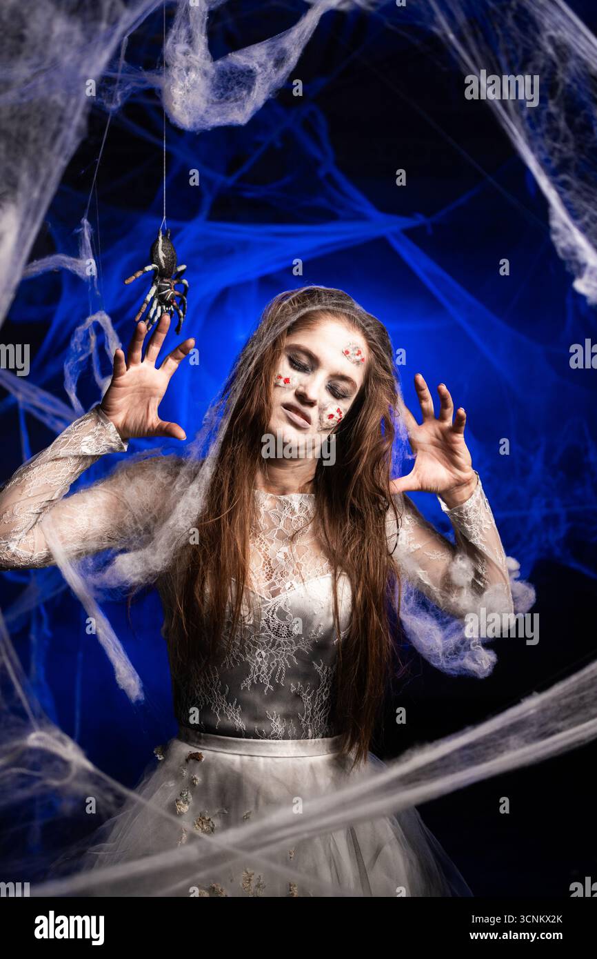 Donna inquietante con orribile trucco zombie di Halloween in abito bianco su sfondo blu circondato da ragnatela. Persona ossessionata dal diavolo. Horro Foto Stock