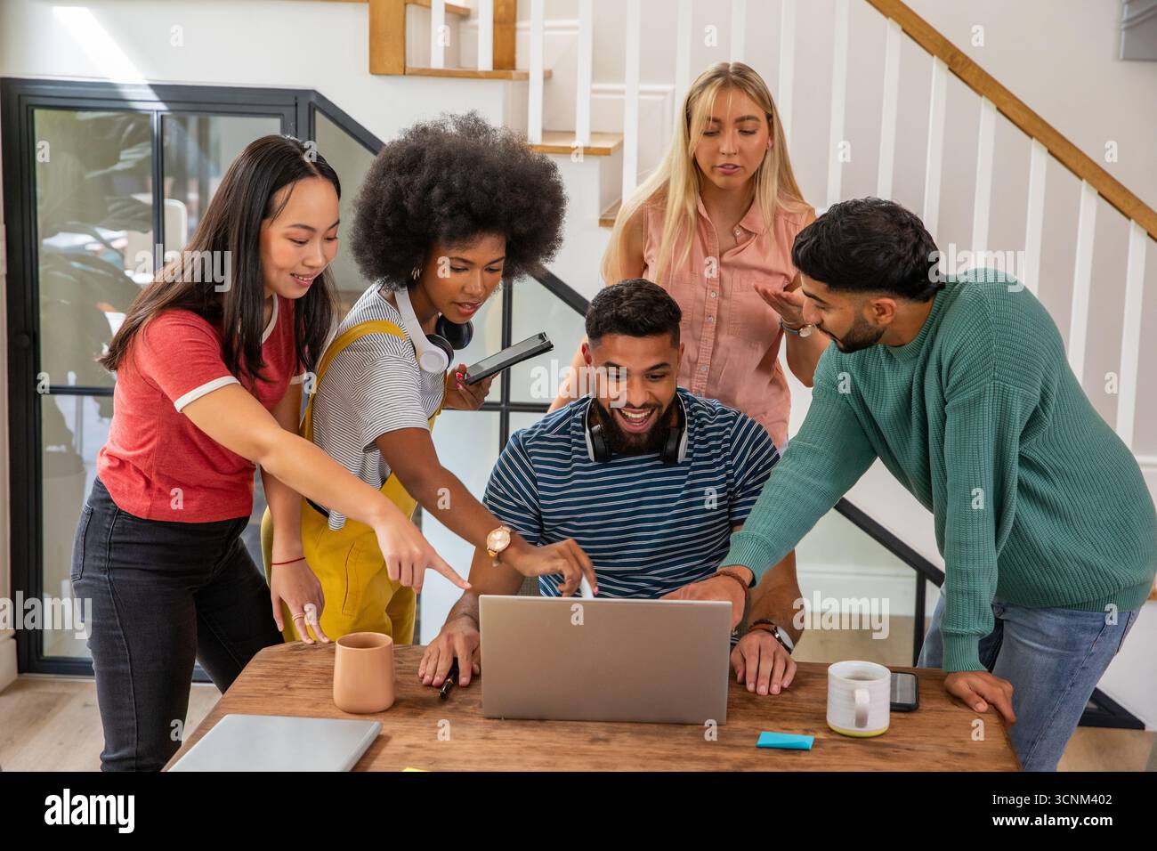 Amici studenti diversi che collaborano intorno al notebook alla base delle scale con tablet e cuffie Foto Stock