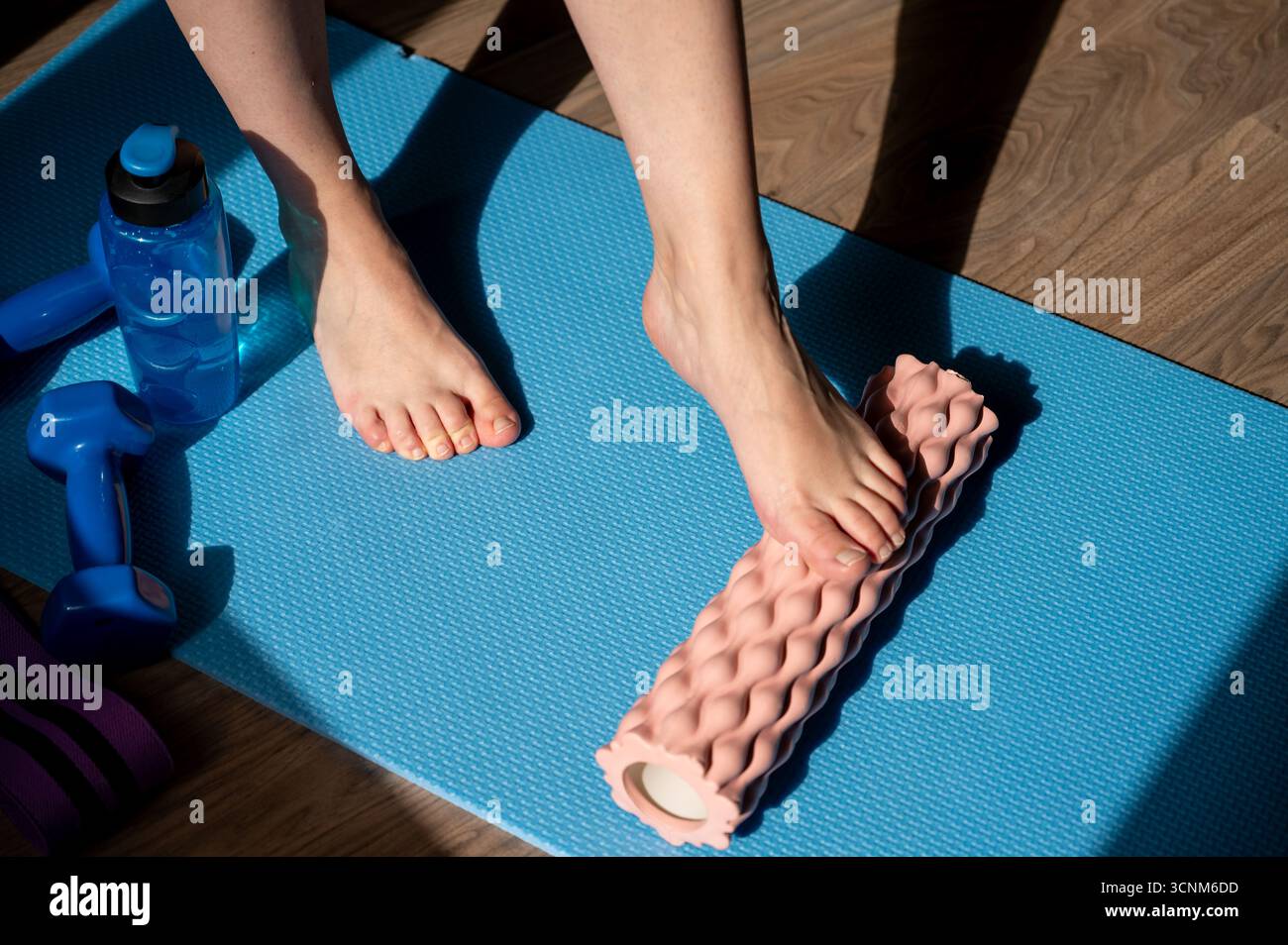 Gambe femminili che eseguono un massaggio a rilascio miofasciale su rullo in schiuma a casa, con manubri e bottiglia d'acqua sul tappetino da yoga, concetto di recupero fitness Foto Stock