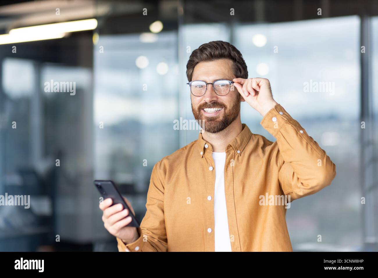 Giovane uomo d'affari in un abbigliamento casual e intelligente sorride con sicurezza mentre si regolano gli occhiali e si tiene in mano uno smartphone, trasmettendo comunicazioni digitali, networking e successo sul luogo di lavoro moderno Foto Stock