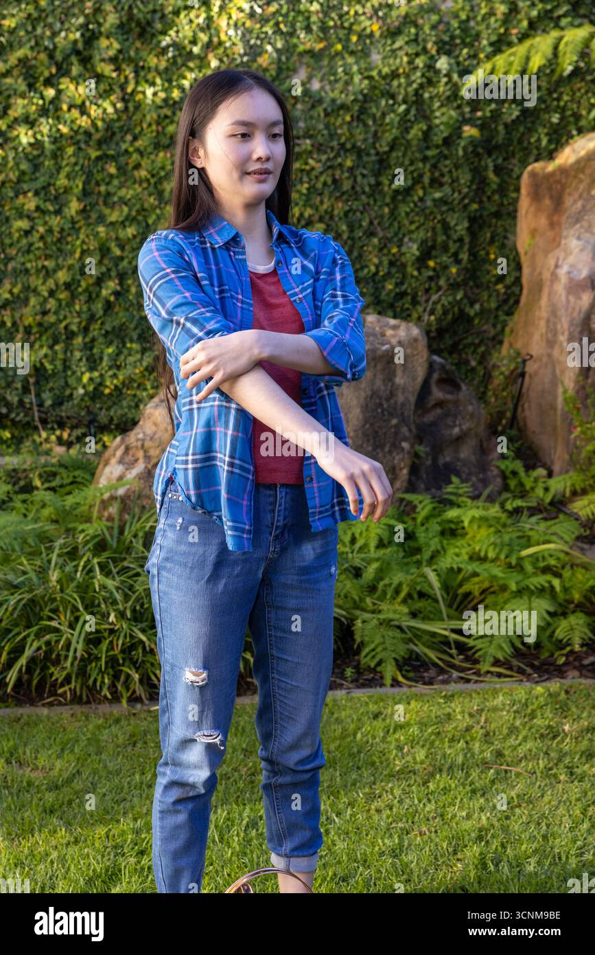 Giovane donna asiatica che regola la manica della camicia sull'erba in giardino tra rocce ornamentali siepe rifilata Foto Stock