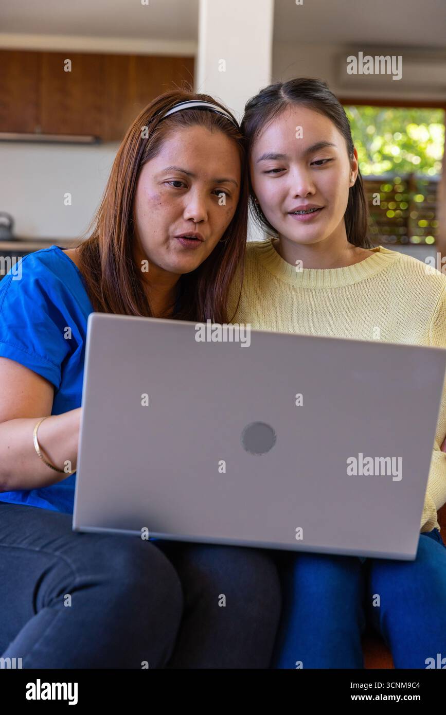 Madre e figlia asiatica sedute sul divano di casa vicino alla cucina imparando insieme sul laptop Foto Stock