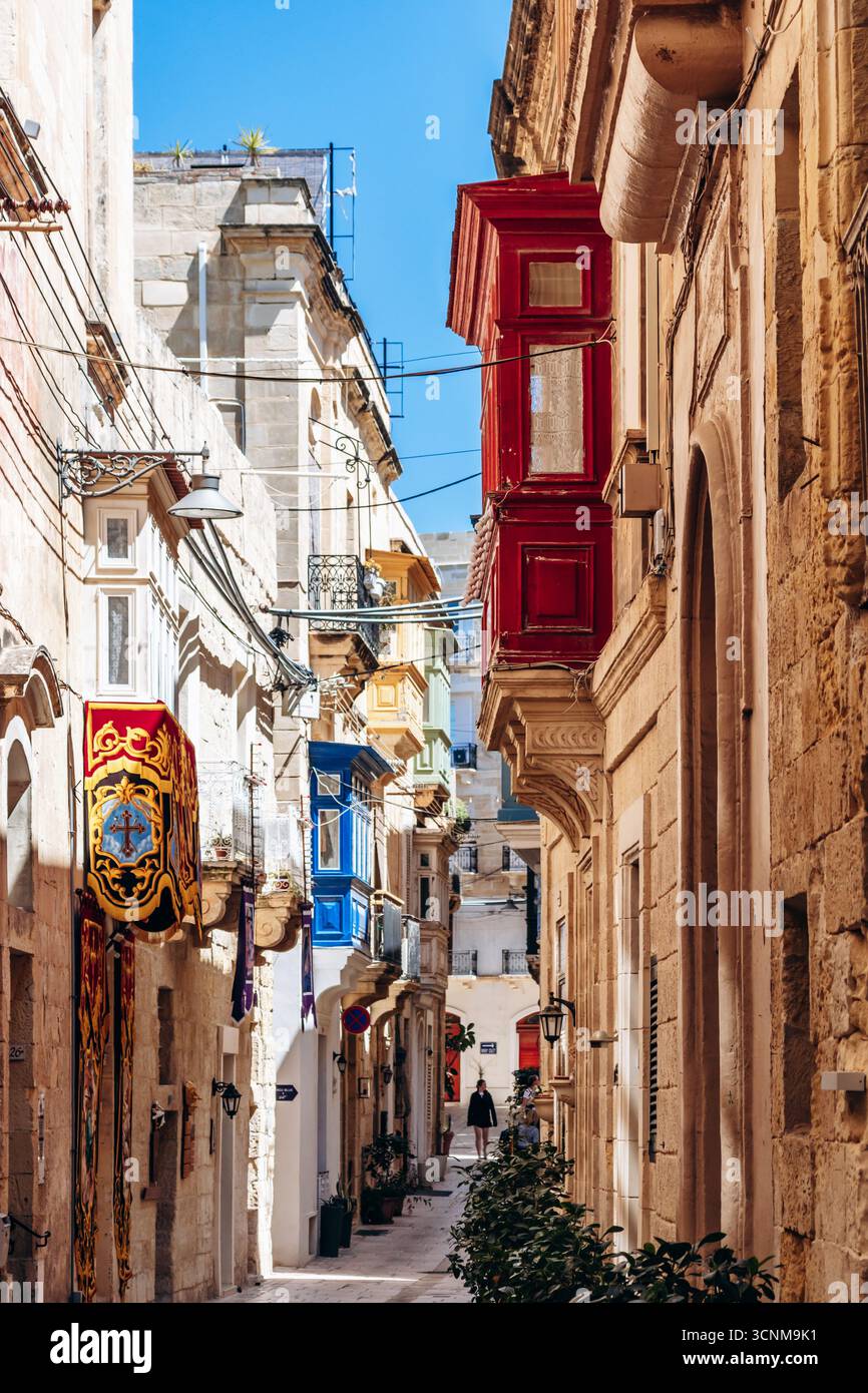BIRGU, MALTA - 11 APRILE 2025: Tradizionale via maltese con balconi in legno colorati, bandiere maltesi rosse e bianche e storico edificio in pietra calcarea Foto Stock