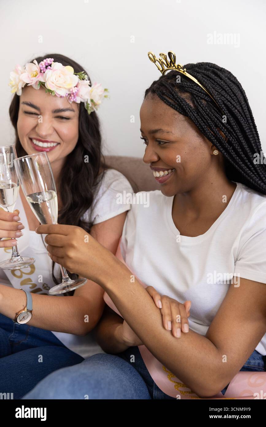 Diverse amiche che indossano l'archetto da sposa con corone floreali che brindono flauti di champagne sul divano Foto Stock