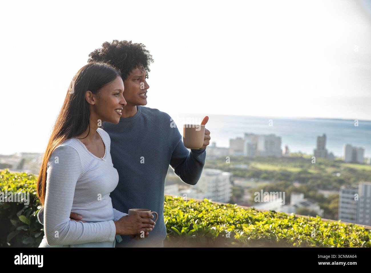 Diverse coppie in piedi sulla terrazza sul tetto vicino al siepe che si affaccia sulla città costiera, tenendo tazze Foto Stock