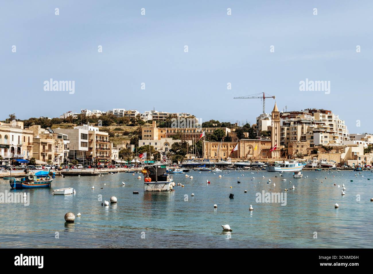 Vista panoramica sul lungomare di Marsaskala con edifici colorati, barche e acque turchesi limpide in un giorno di primavera soleggiato, Malta, Europa Foto Stock