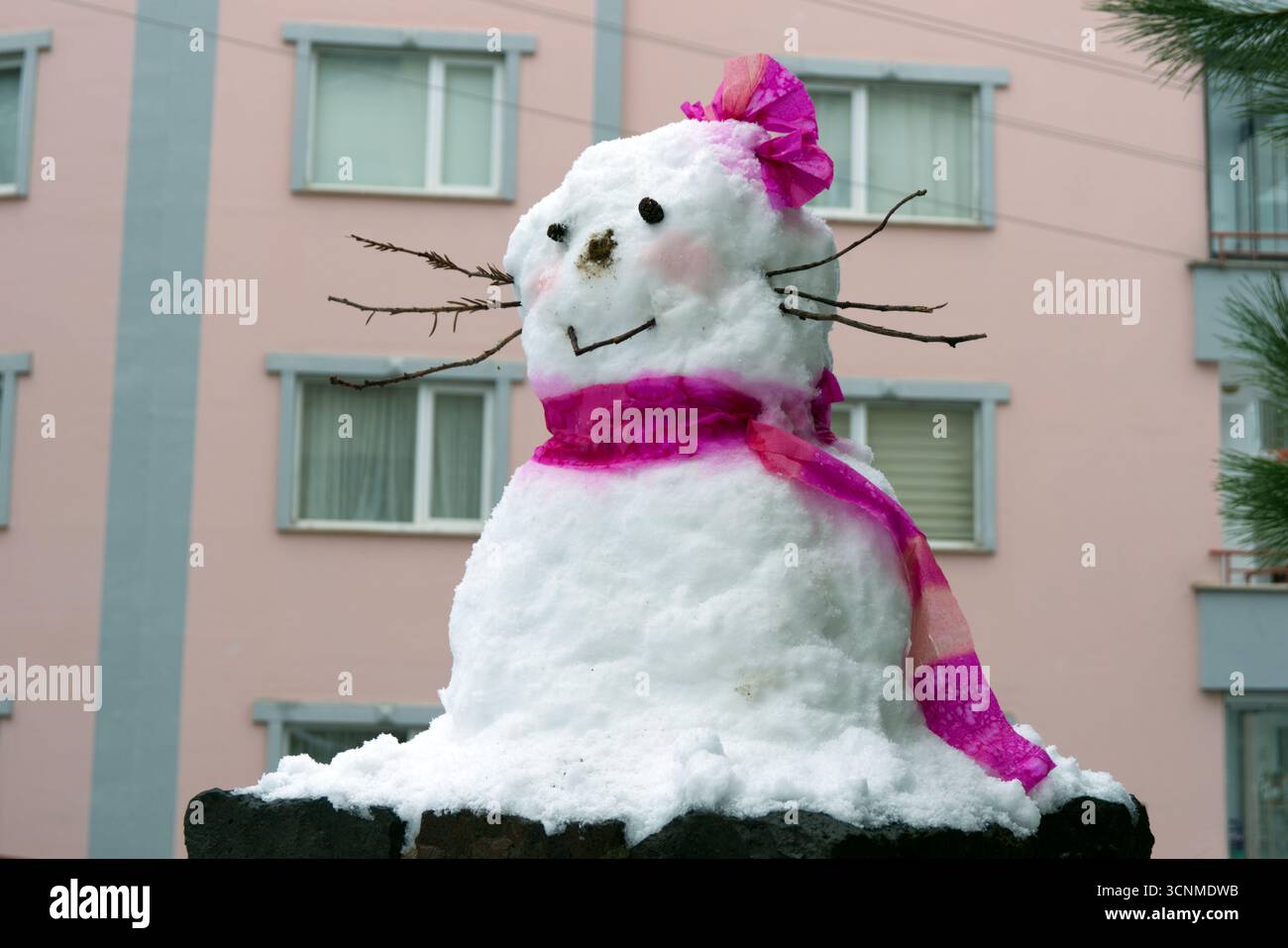 Grazioso pupazzo di neve con sciarpa rosa e arco. Un pupazzo di neve disegnato con sciarpa rosa, arco e bastoncini simili a un frullatore, che donano un aspetto giocoso e carino. Foto Stock