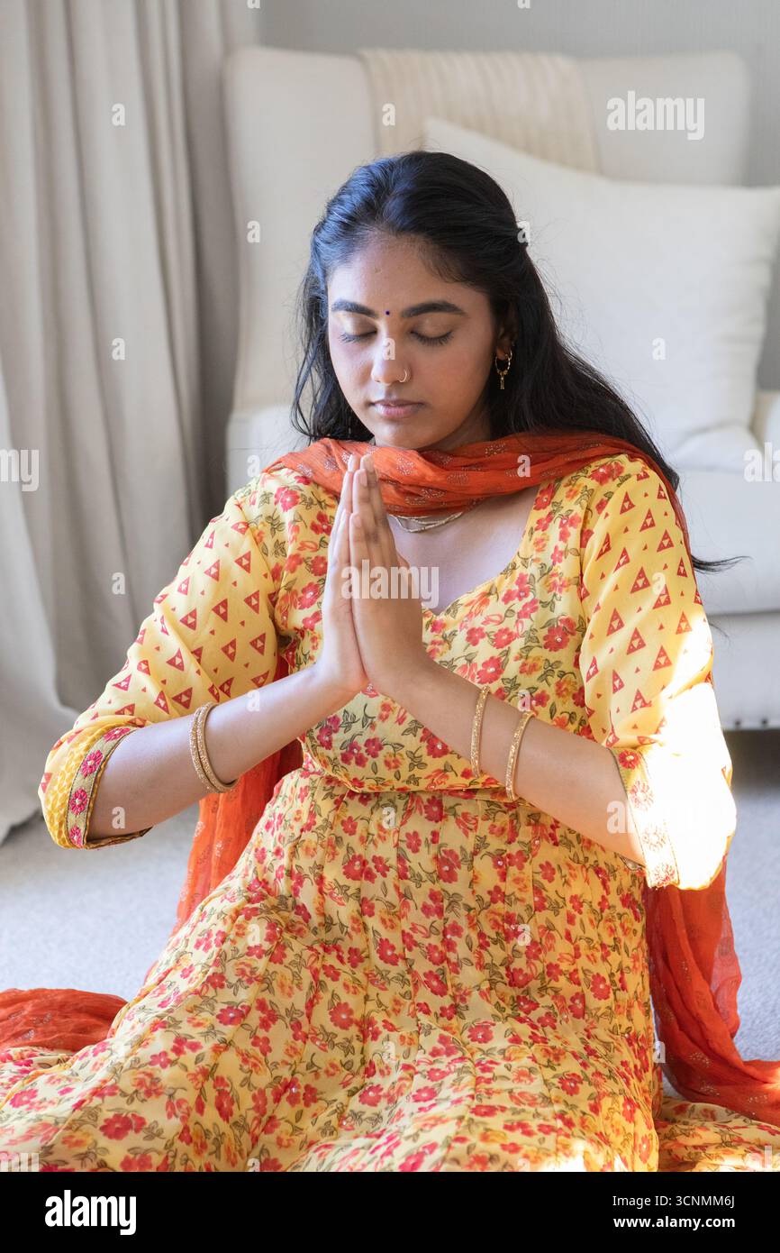 Donna indiana in kurta giallo che prega con le palme premute al petto su tappeto nella camera da letto di casa Foto Stock