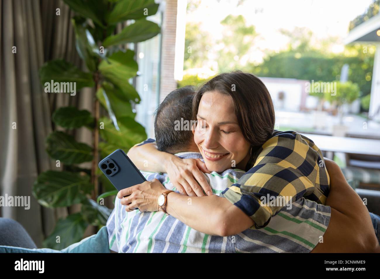 Diverse coppie senior che abbracciano il divano tramite porta scorrevole in vetro con smartphone e impianto in vaso Foto Stock