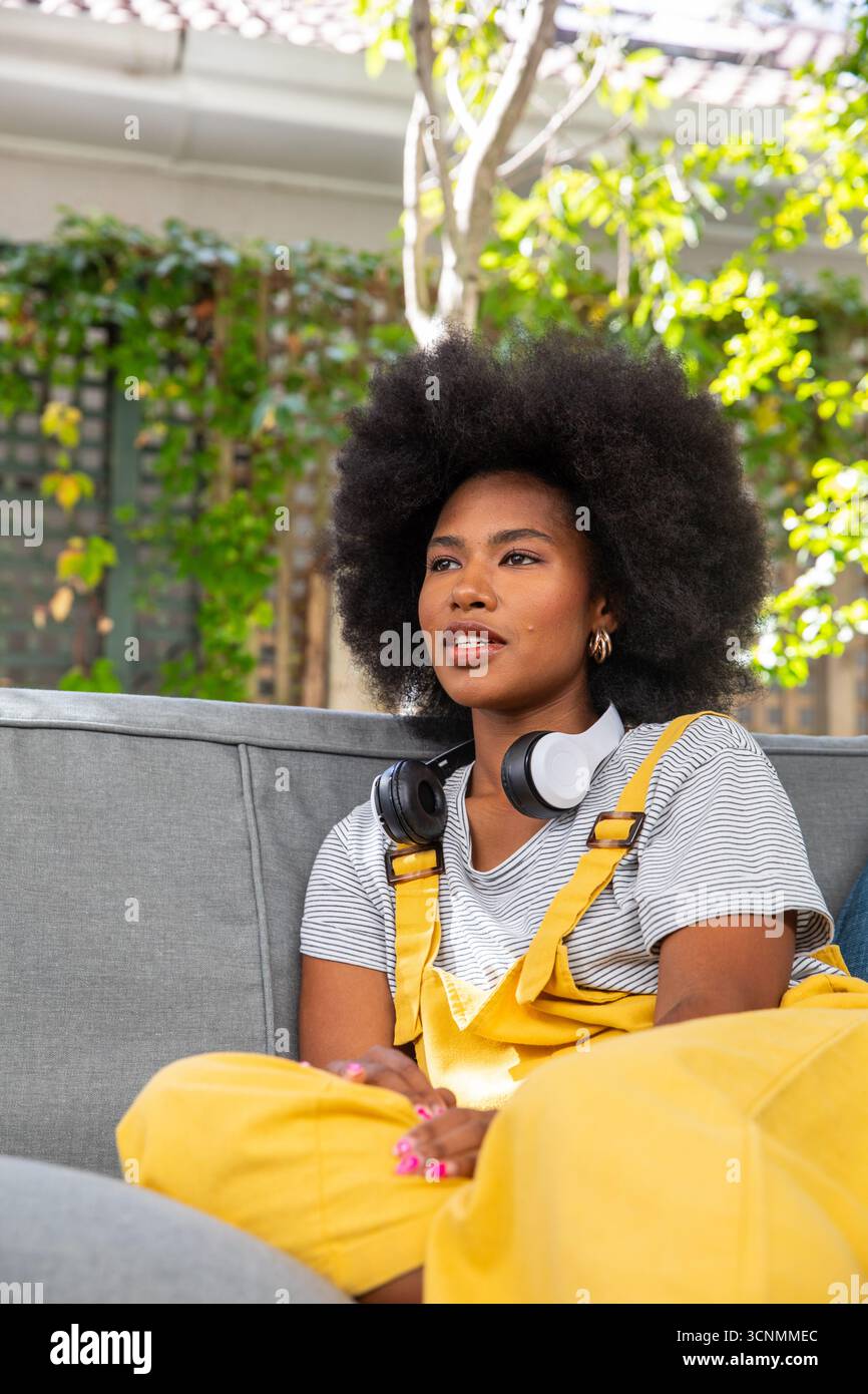 Donna afroamericana seduta sul divano nel patio del giardino con le cuffie Foto Stock