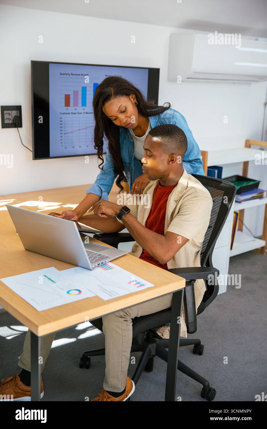 Colleghi afroamericani che collaborano su un laptop argentato alla scrivania dell'ufficio, analizzando le carte Foto Stock