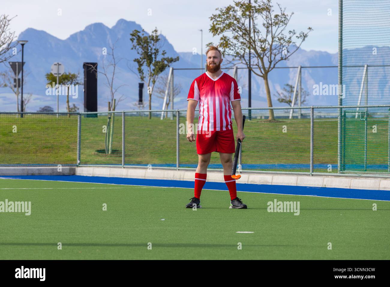 Uomo in piedi su un tappeto erboso sintetico nel complesso sportivo con bastone da hockey su una recinzione metallica a rete Foto Stock