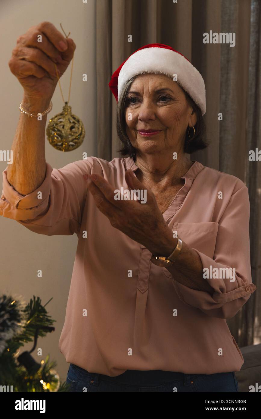Donna anziana che tiene in mano la pallina di Natale d'oro accanto all'albero di Natale decorato a casa nel cappello di Babbo Natale Foto Stock