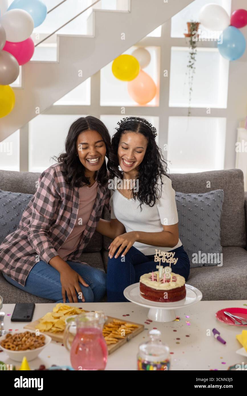 Mamma e figlia adolescente diverse sedute sul divano accanto alle scale per festeggiare il compleanno con palloncini per torte Foto Stock