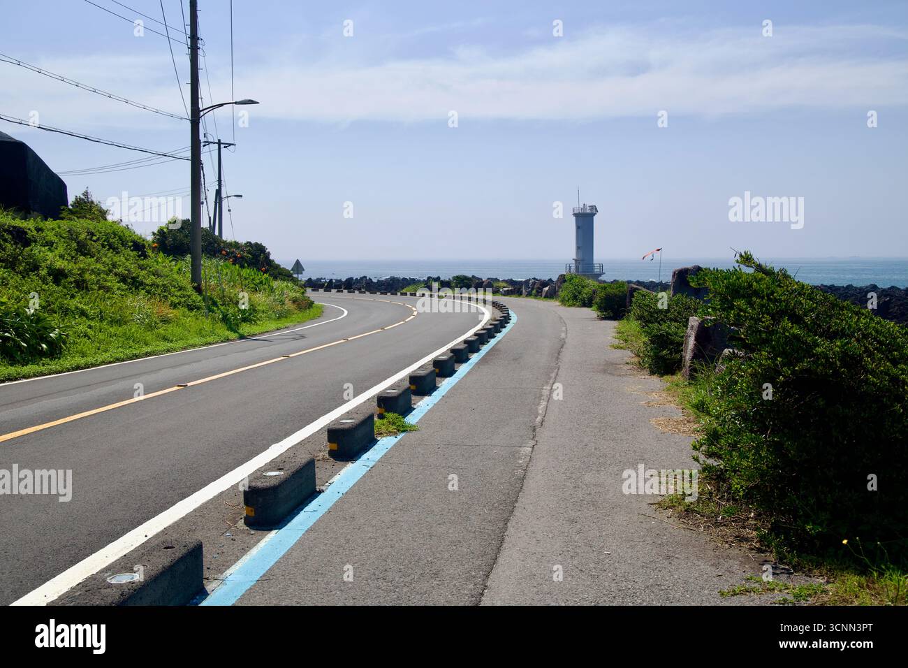 Una curva A S sulla strada costiera si avvicina a un faro bianco vicino a Sinheung-ri, con una pista ciclabile separata e rocce basaltiche lungo la costa. Foto Stock