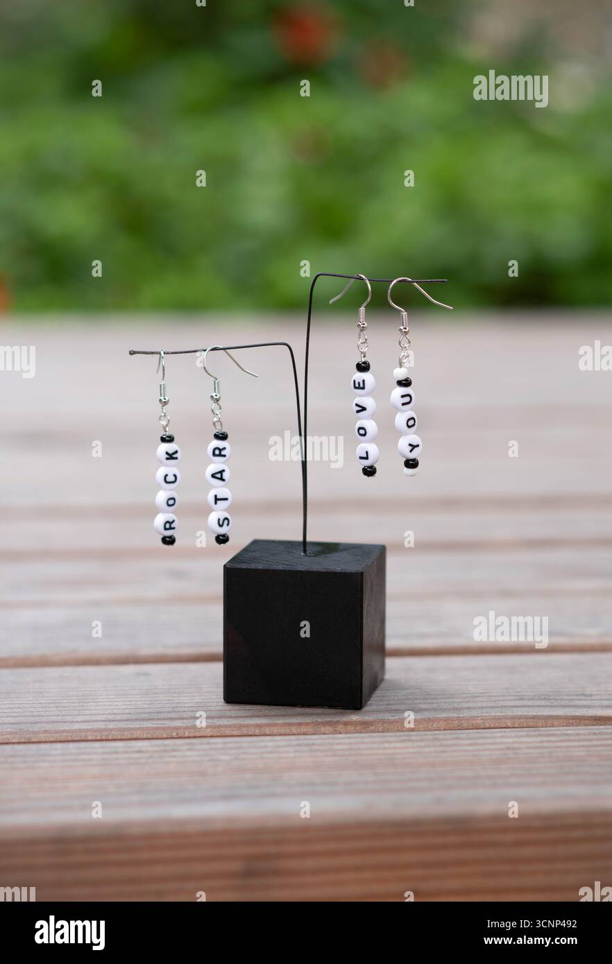 Orecchini fatti a mano con perline dell'alfabeto bianco e nero che scrivono "ROCK STAR" e "LOVE YOU", visualizzati su un piedistallo nero minimalista su sfondo in legno. Foto Stock