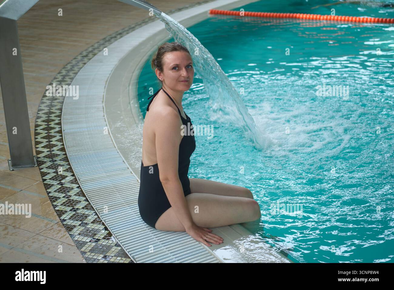 Donna in costume da bagno nero siede a bordo piscina con il flusso d'acqua che scorre. Centro benessere e benessere con terapia acquatica. Foto Stock