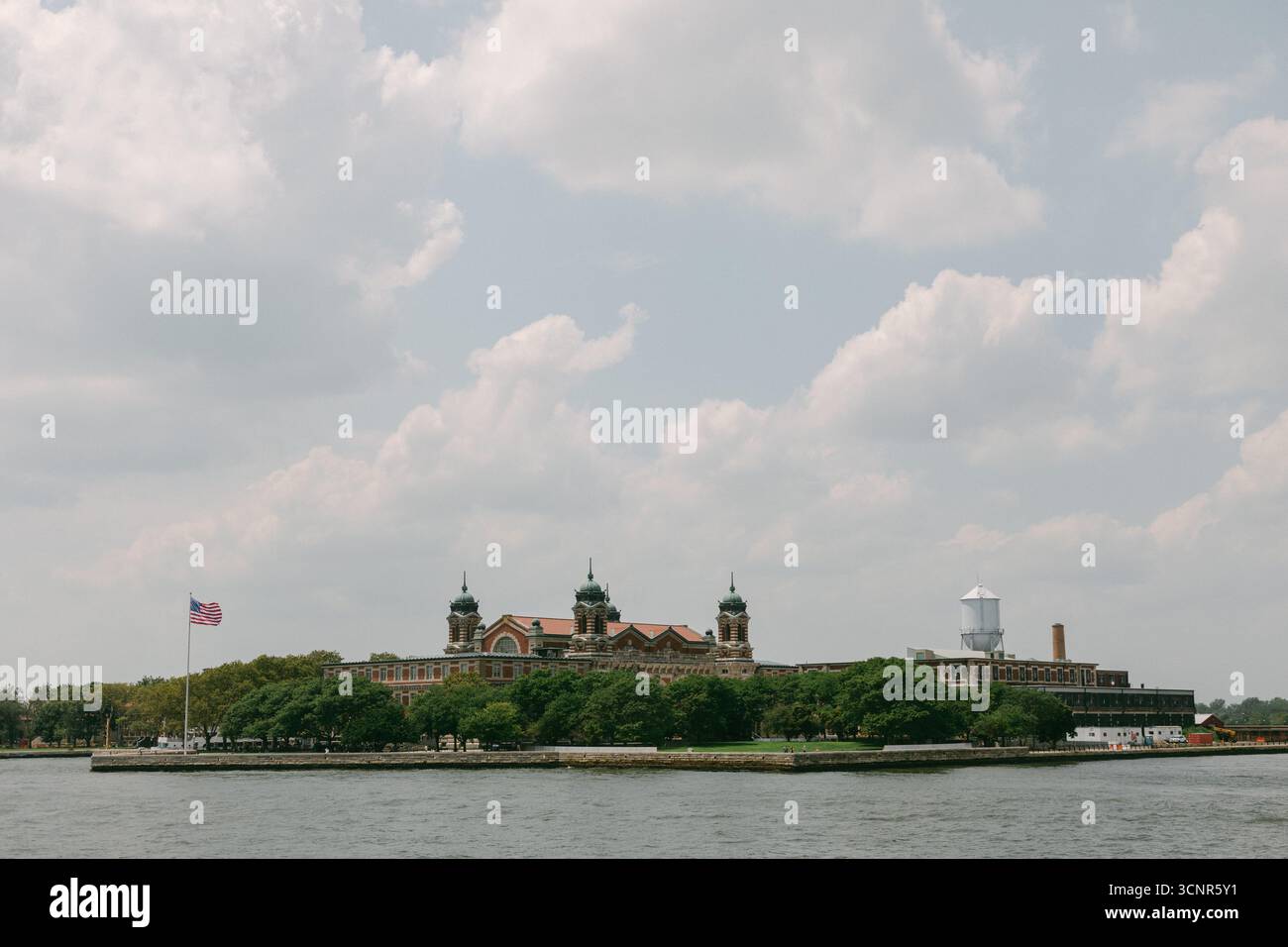 Vista di Ellis Island con i suoi edifici storici e la bandiera americana, sullo sfondo di nuvole. Foto Stock