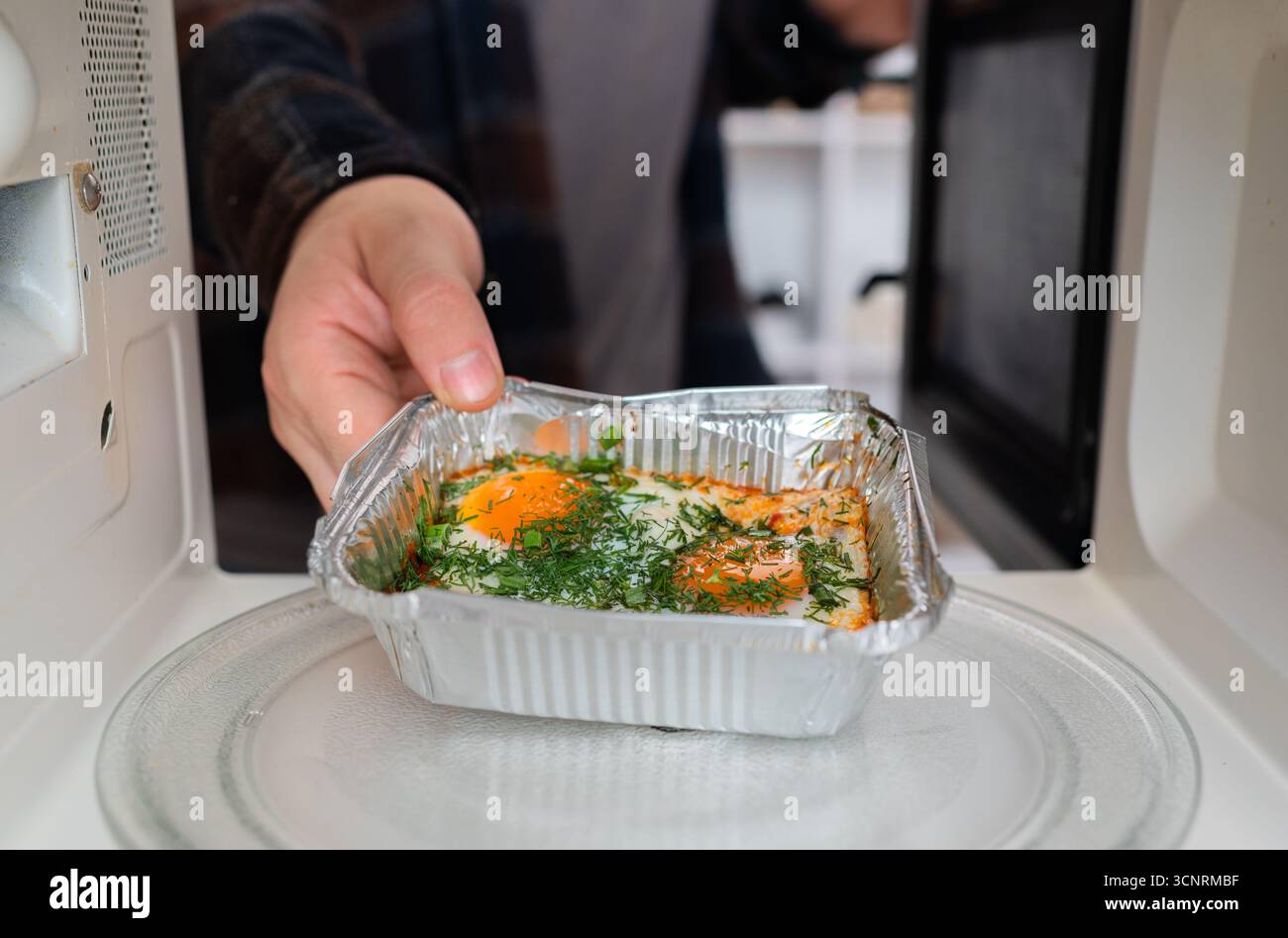 La mano di una persona posiziona un contenitore per asporto in alluminio con il cibo in un microonde, un errore comune in cucina e un problema di sicurezza Foto Stock