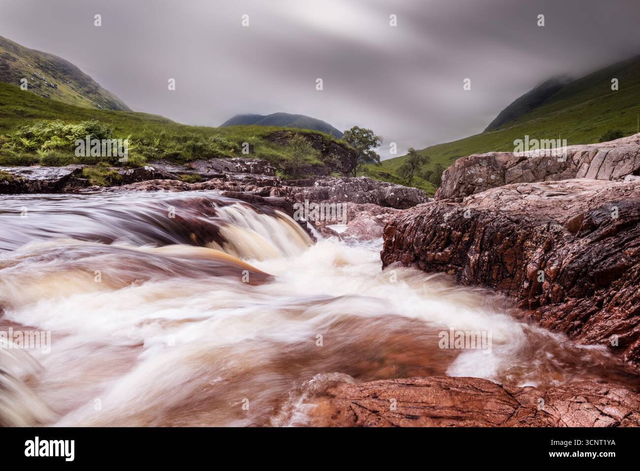 La cascata di fiume selvaggia che scorre su rocce aspre nel suggestivo paesaggio delle Highlands scozzesi con cielo nebbioso e lussureggianti colline verdi Foto Stock
