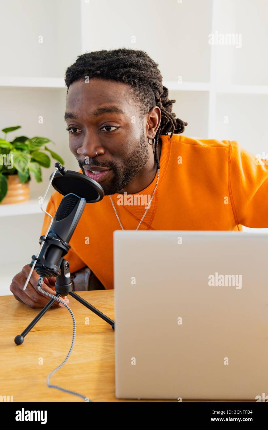 Uomo afroamericano che si appoggia in avanti alla scrivania dello studio e parla nel microfono a condensatore con un computer portatile Foto Stock