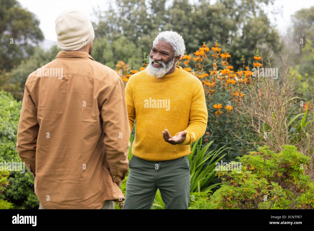 Amici maschi anziani in piedi in un giardino lussureggiante con arbusti in fiore arancione e maglione a maglia di senape Foto Stock
