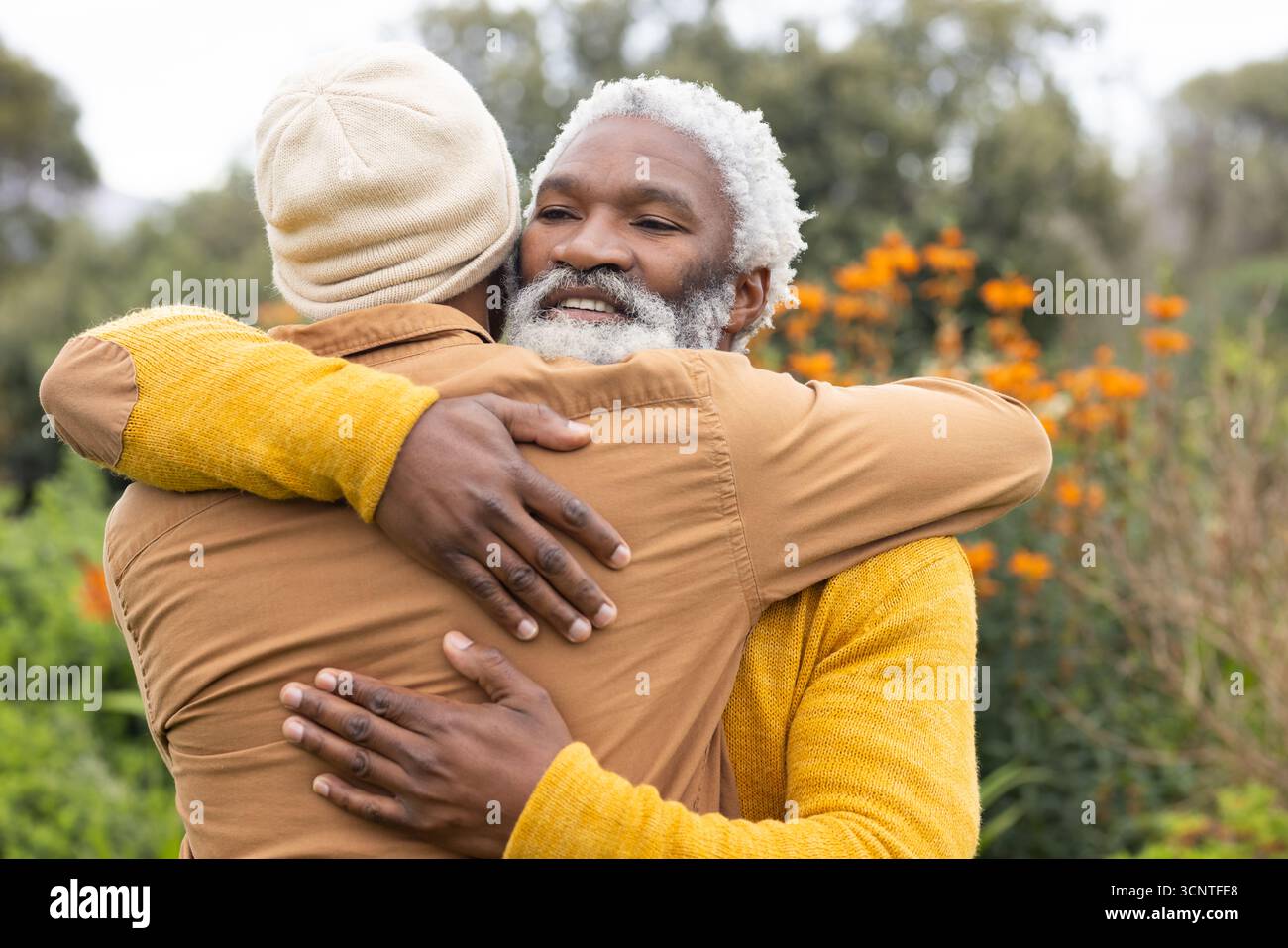 Padre e figlio afroamericani abbracciati in giardino tra arbusti arancioni con maglione in maglia gialla Foto Stock