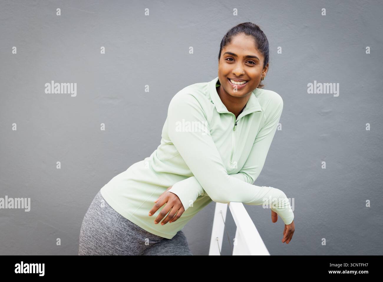 Donna dell'Asia meridionale in abbigliamento atletico appoggiata su ringhiere in metallo bianco sulla terrazza sorridente alla macchina fotografica Foto Stock