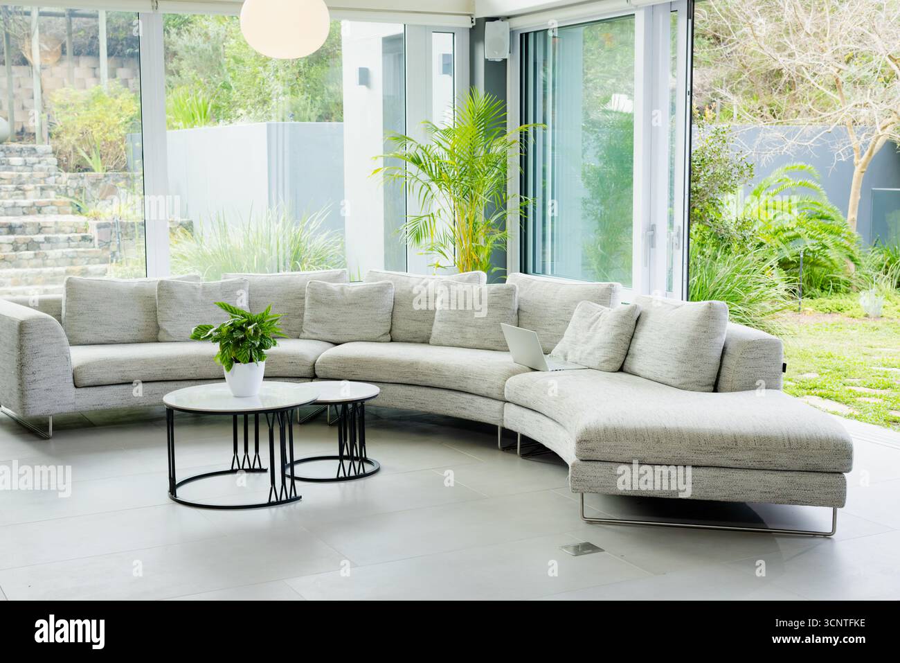 Divano curvo che circonda tavolini da caffè con pianta e computer portatile, pareti in vetro che rivelano palme da giardino Foto Stock