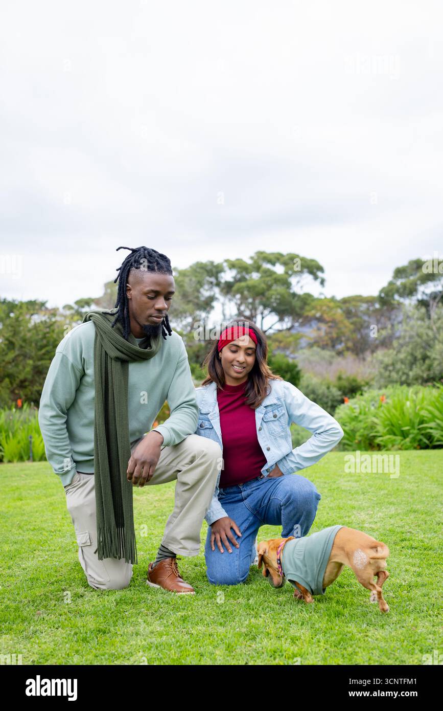 Diverse coppie si inginocchiano nel parco con sciarpa d'oliva e fascia rossa che accarezzano il cane maglione verde abbronzante Foto Stock