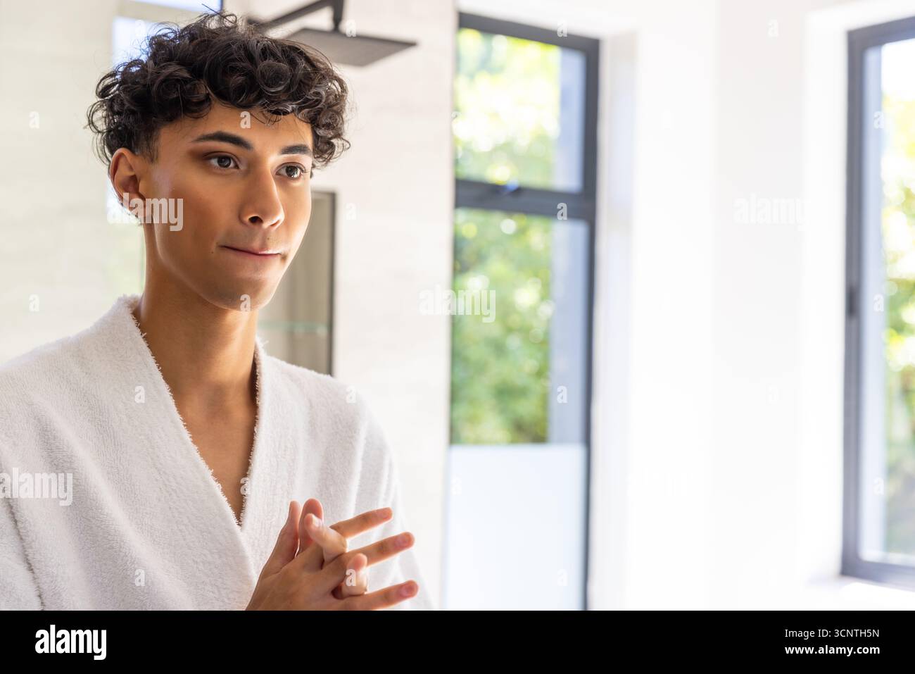 Uomo mediorientale che applica prodotti per la cura della pelle nel bagno con accappatoio e finestre incorniciate Foto Stock