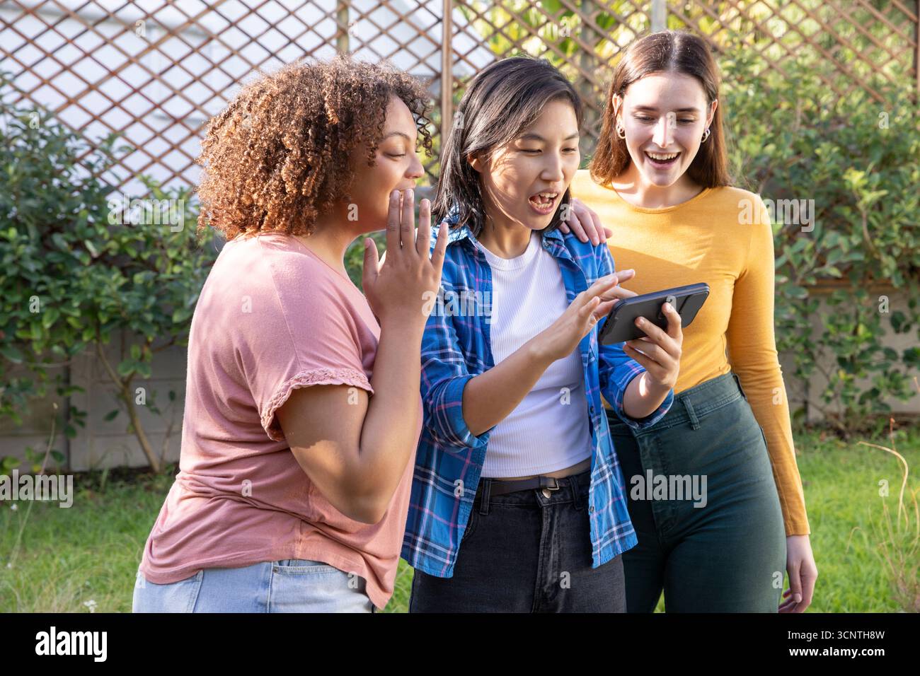 Diverse amiche che si appoggiano e visualizzano un tablet insieme nel giardino soleggiato del cortile Foto Stock