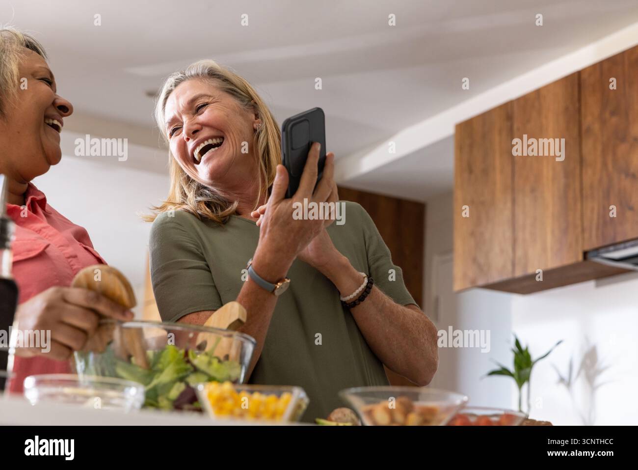 Le amiche ridono, gettano l'insalata con un cucchiaio e controllano lo smartphone al banco della cucina Foto Stock