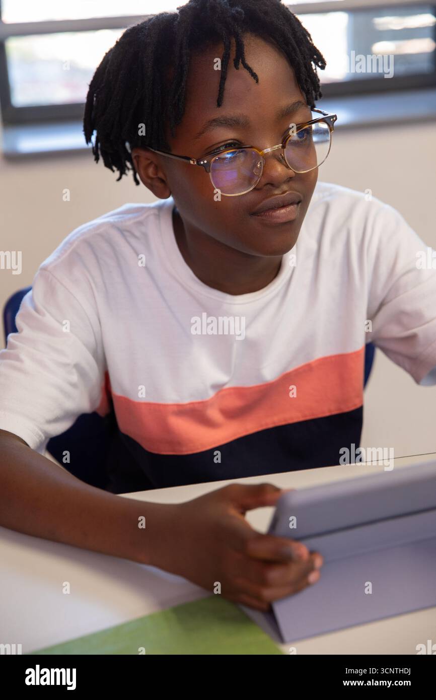 Studentessa afroamericana seduta alla scrivania in classe con occhiali da vista e tablet in mano Foto Stock