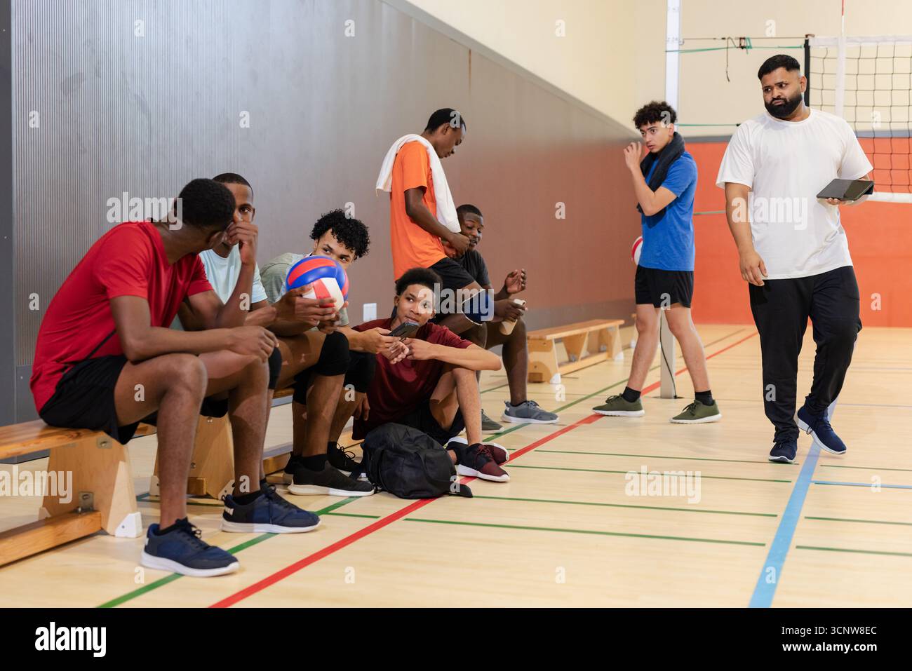 Diverse squadre di pallavolo maschile su panchina di legno che tengono pallavolo in palestra mentre l'allenatore cammina con TAB Foto Stock