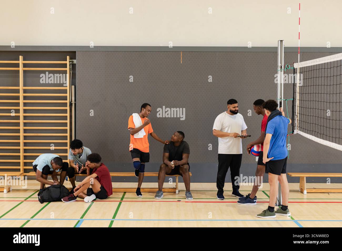 Diversi compagni di squadra maschili che fanno una pausa in campo intorno a borse sportive e pallavolo vicino alla rete Foto Stock