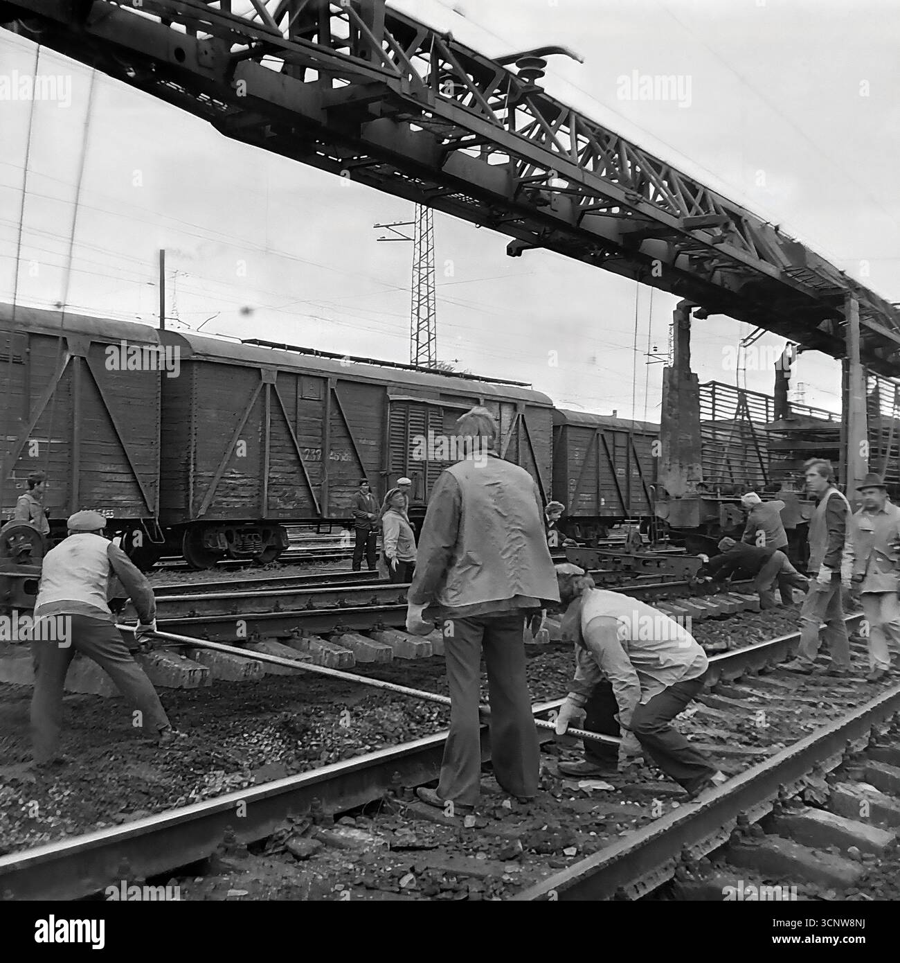 Una fotografia d'archivio degli anni '1970 cattura un momento di precisione tecnica durante un progetto di costruzione ferroviaria a Sloviansk, RSS Ucraina. Un team di operai ferroviari sovietici utilizza un lungo strumento di misurazione per controllare lo scartamento e l'allineamento di una nuova pista, mentre un supervisore supervisiona il processo critico di controllo della qualità. Ambientato in uno scenario industriale di autocarri e gru a cavalletto, questa immagine mette in evidenza l'ingegneria e il lavoro di squadra necessari per costruire le infrastrutture dell'URSS Foto Stock