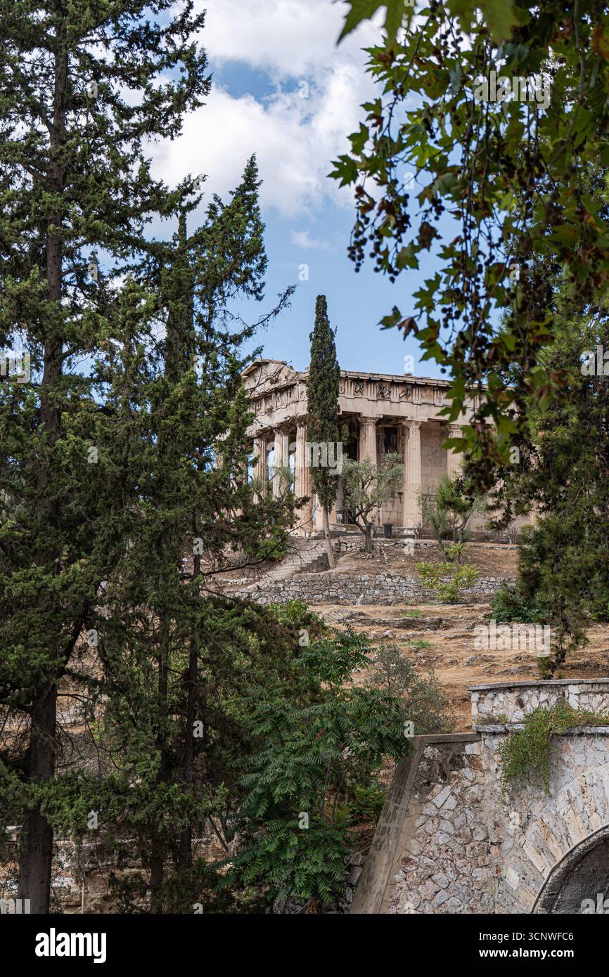 L'antico Tempio di Efesto ad Atene, parzialmente nascosto da vegetazione lussureggiante e alberi, mostra l'architettura classica sotto un cielo limpido e blu, simb Foto Stock
