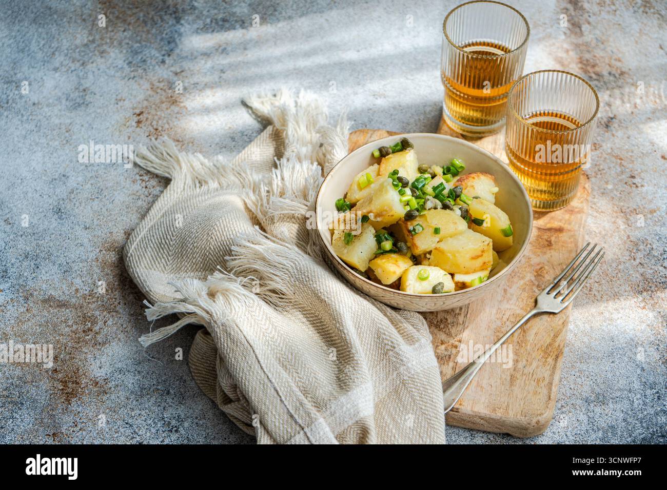 Una deliziosa insalata di patate greche guarnita con capperi e scalpelli a fette, servita in un'elegante ciotola su una tavola di legno con due bicchieri di tè e un Foto Stock