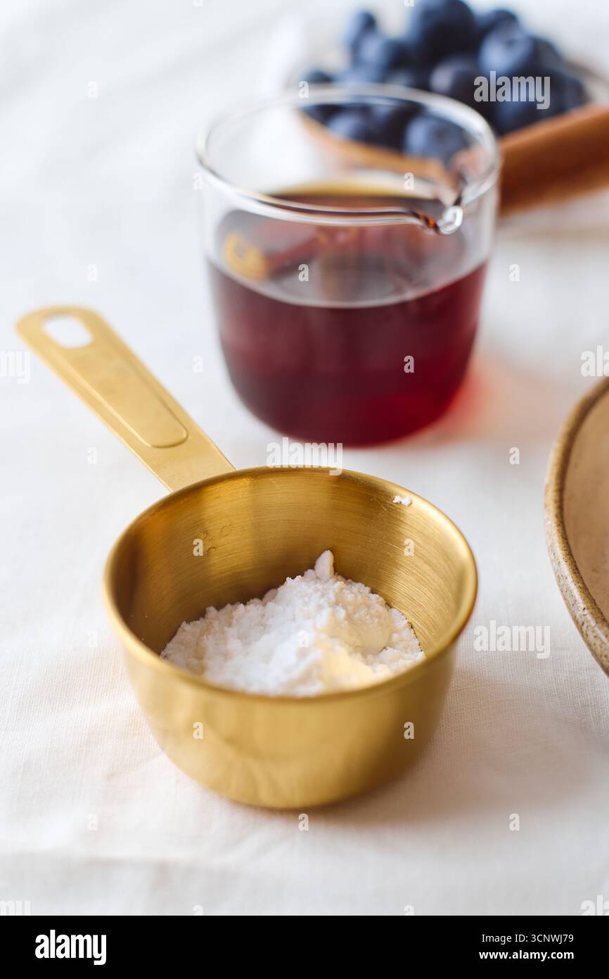 Un dosatore di ottone riempito di farina si trova accanto a un bicchiere di succo di bacca e mirtilli freschi su una tovaglia bianca, pronto per una deliziosa cucina Foto Stock