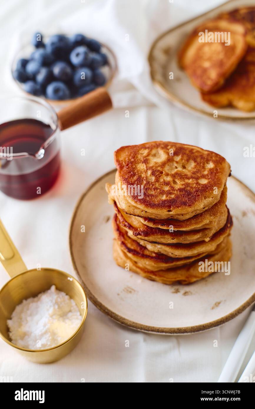 Una pila di soffici pancake dorati si trova su un piatto, accompagnati da mirtilli freschi e una piccola caraffa di sciroppo. Un dosatore con farina aggiunge un tocco Foto Stock