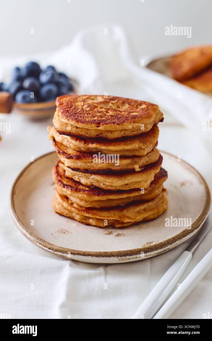 Una pila di frittelle dorate si trova su un piatto rustico in ceramica. Lo sfondo presenta mirtilli freschi che aggiungono un tocco di colore e freschezza Foto Stock